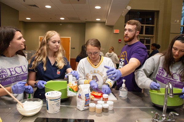 A bearded man in a purple T-shirt talks to a group of women preparing food in a large meeting room.